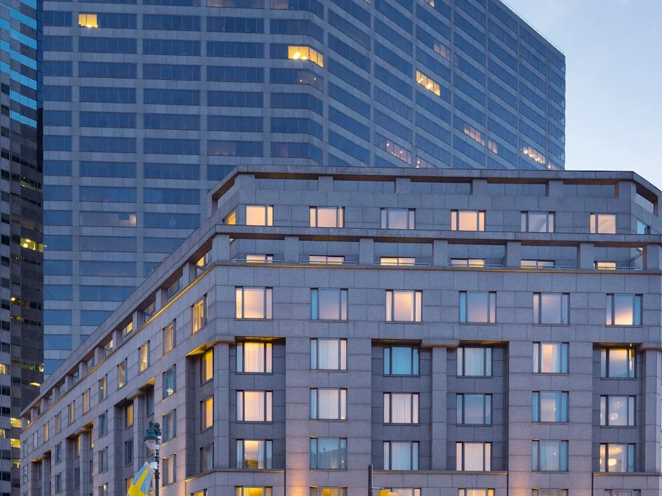 A gray stone hotel building at dusk in front of a taller glass tower