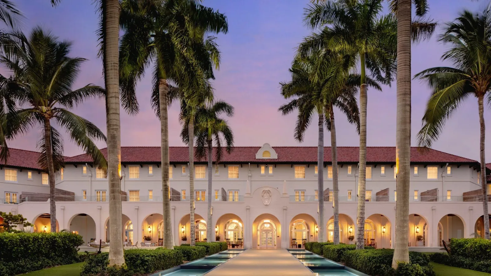 Front view of a Mediterranean-style hotel with an arched colonnade, warm-lit windows, and tall palm trees at sunset.