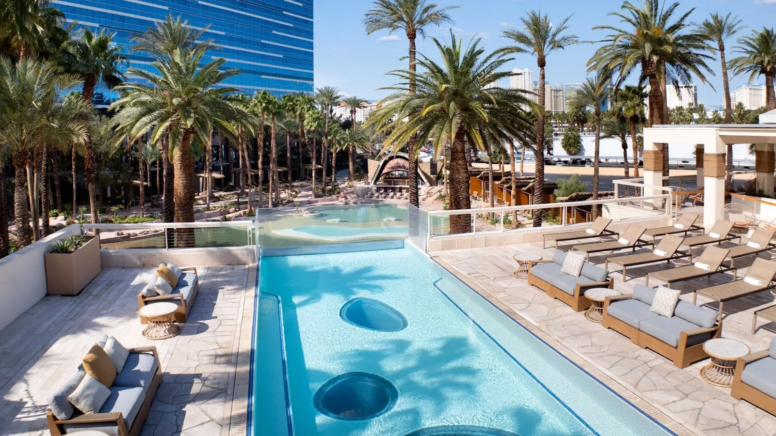 Luxurious pool deck with palm trees, lounge chairs, and a clear blue pool against a cityscape backdrop.